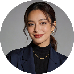 Smiling woman with dark hair in a ponytail, wearing gold hoop earrings, a gold necklace, a black turtleneck, and a navy blazer against a gray background.